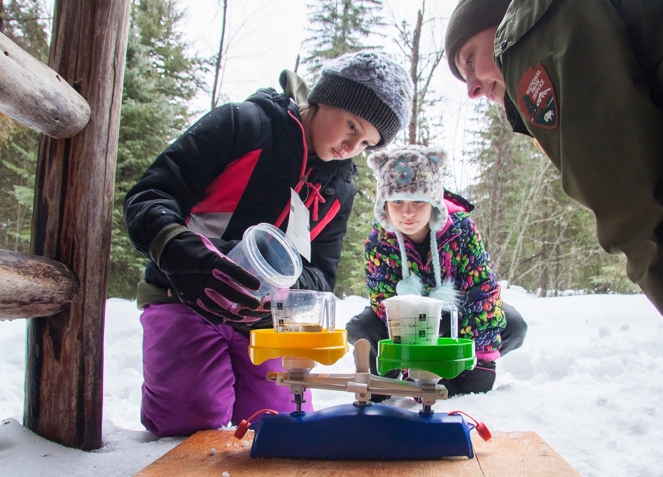 A ranger and two students use a scale to measure cups of snow and water.