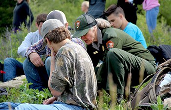 Uniformed employee crouches by students with notebooks in field