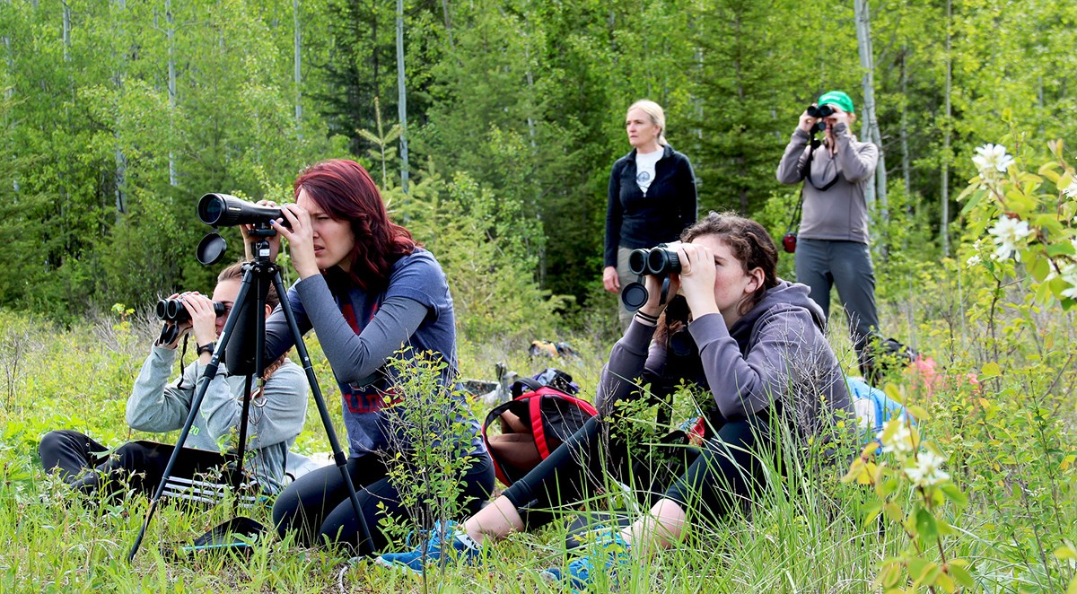 group of students look through binoculars while sitting on ground, with teachers standing behind group of students look through binoculars while sitting on ground, with teachers standing behind