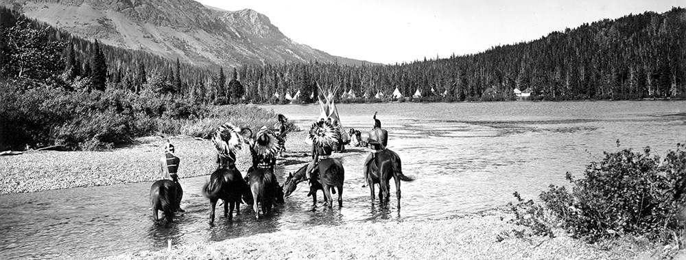 Five Native Americans on horseback cross mountain stream toward camp