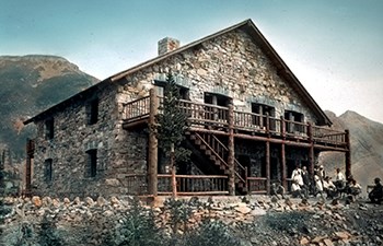 tinted historic slide of stone building with large wooden porch and people in period dress seated at base