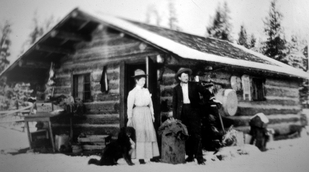 woman, man, and dog in front of rustic log cabin