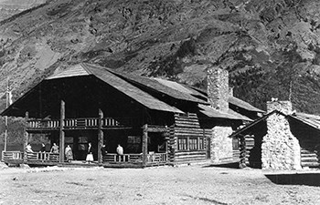 historic image of people on porch of large 2 story log building with smaller log cabin beside