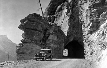 black and white photo of early 1900s car by tunnel in rocky cliff