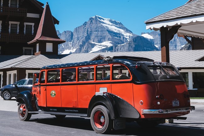 An old bright red bus is parked in front of a hotel lobby with mountains in the background.