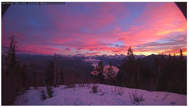 A pink, blue, and orange partly cloudy sky at sunrise glows above snowy mountains, a large lake, and forests.