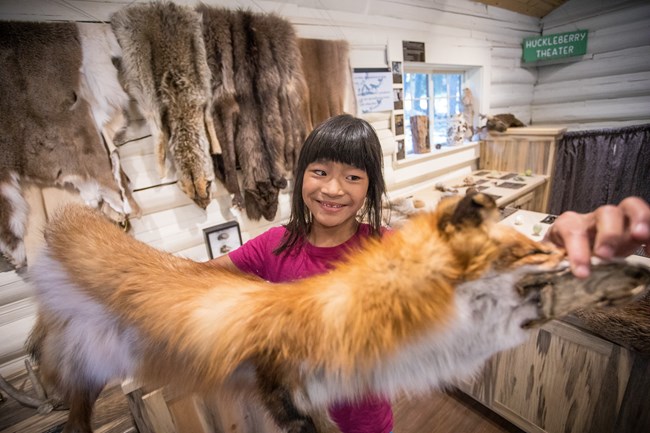 A young, smiling girl holds a fox pelt up to the camera.