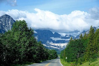 A road leads toward snowy, icy, mountains with trees nearby.