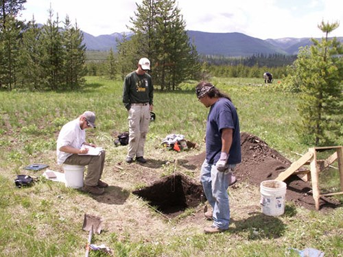 Two men stand over square hole in field while a seated man takes notes