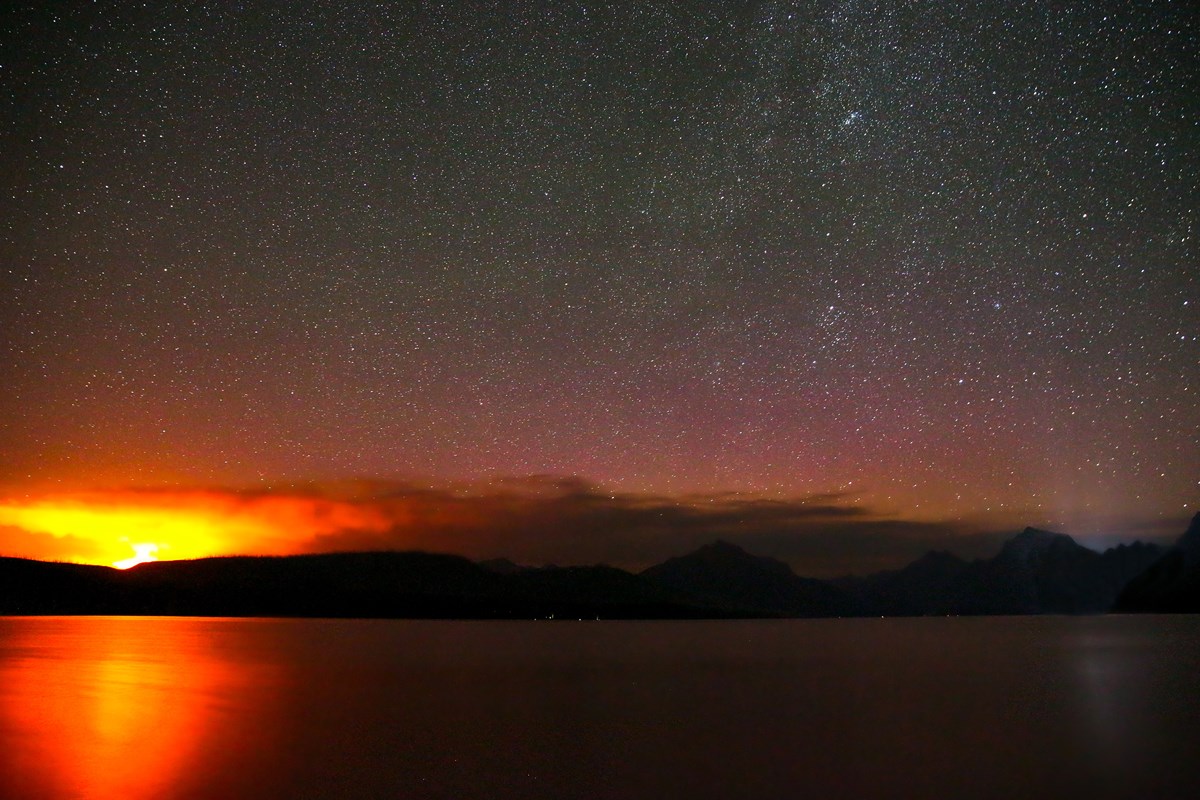 An orange-yellow glow hovers above silhouetted mountains standing below a star-laden sky.