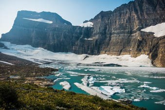 Grass leads down the a icy blue lake with a glacier in the background below a rocky mountain.