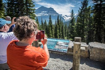 People photograph a sign with their phones. Trees, mountains, and glaciers, are in the background.