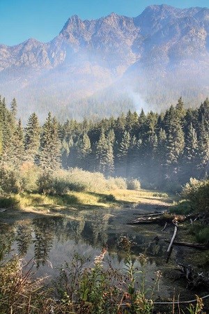 Smoke rises from distant trees standing behind a small pond. A looming mountain serves as a backdrop. Smoke rises from distant trees standing behind a small pond. A looming mountain serves as a backdrop.