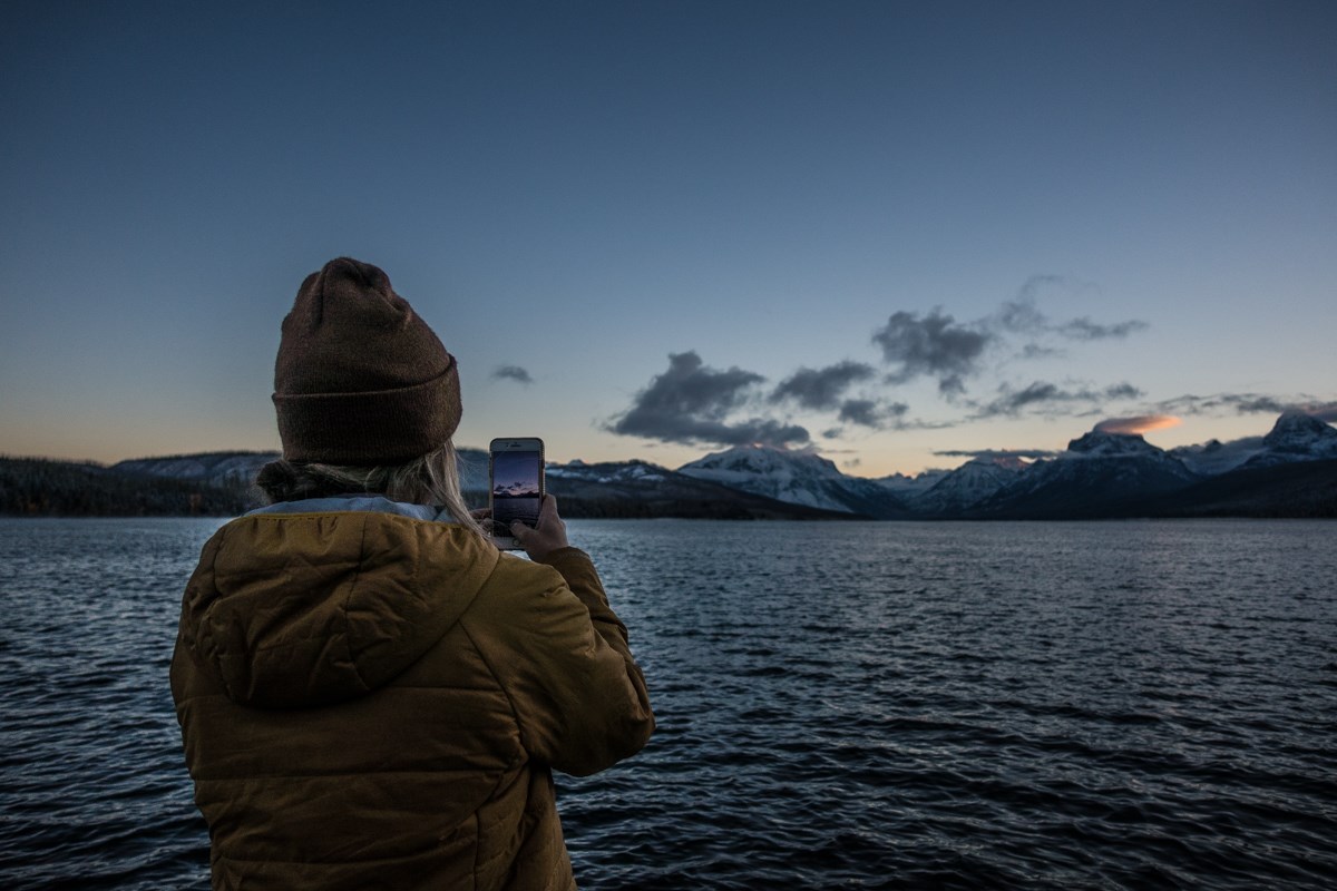 A person in a coat and hat photographs sunrise over mountains with a lake in the foreground.