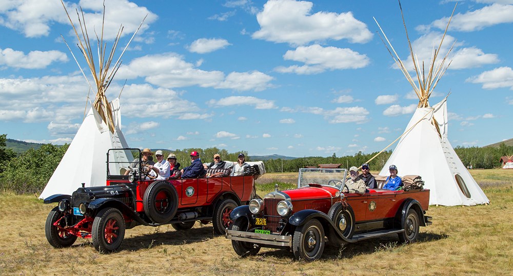 roofless historic red touring cars with seated passengers in front of tipis