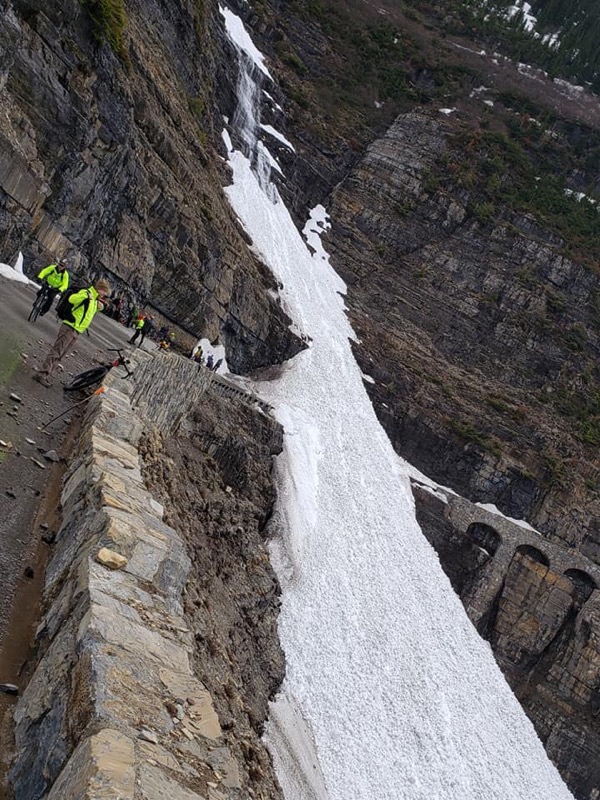 Cyclists stop on narrow mountain road and look at large flow of avalanche debris blocking road.