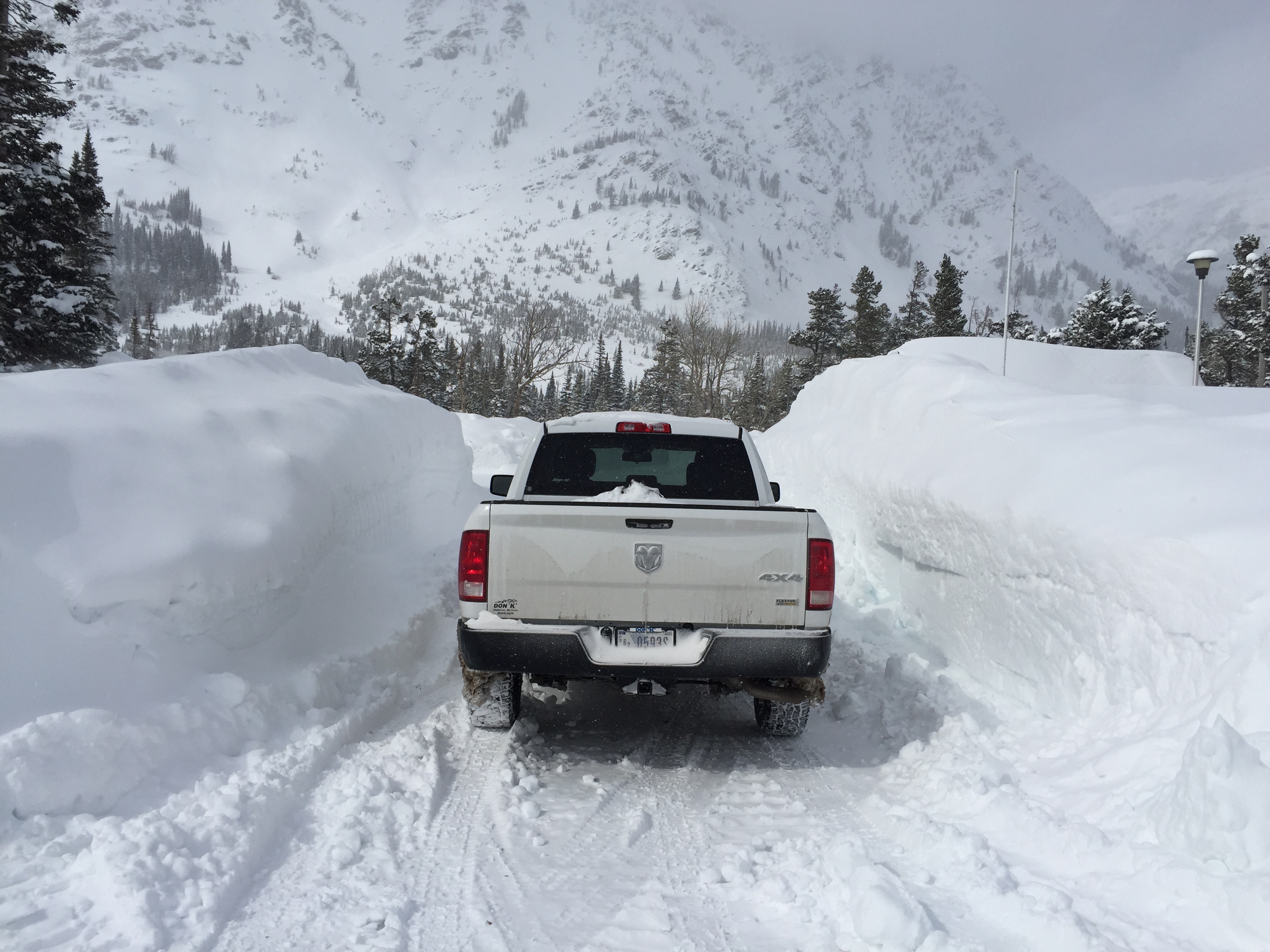 A white truck drives down a snow covered single lane road that is snow packed. A mountain is in the distance. Snow banks tower on either side up to the top of the truck.