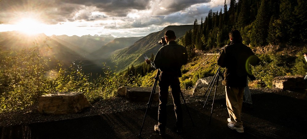 two photographers with tripods face a sunset over mountains