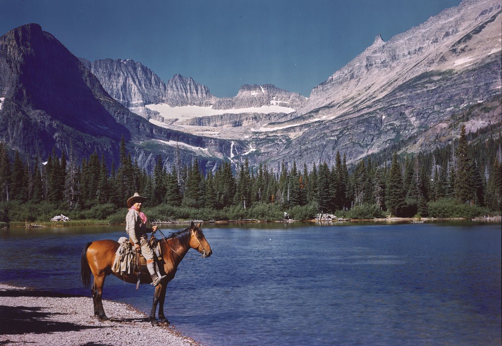 A single rider on horseback poses next to a deep blue lake with mountains and a forest in the background.