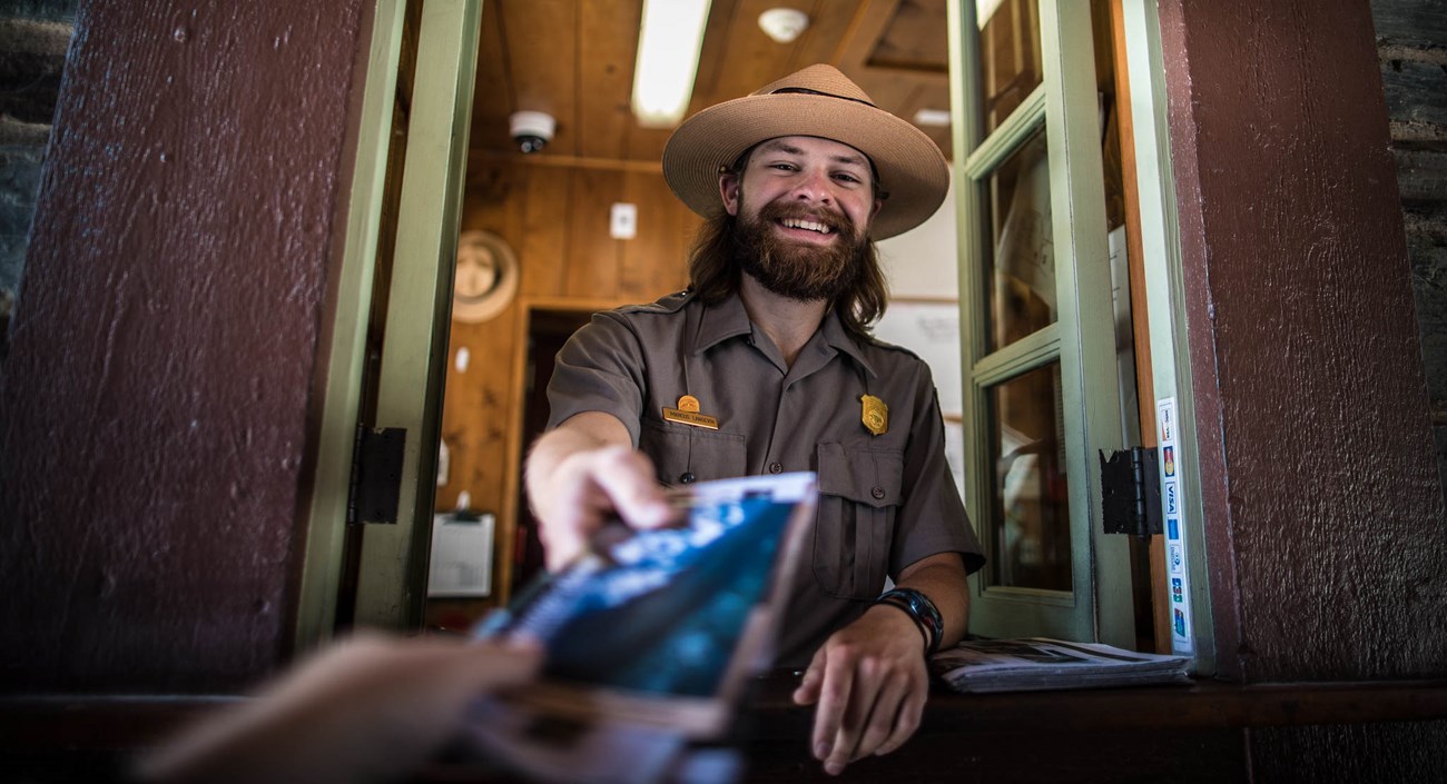 A ranger with a beard wearing a park uniform smiles and hands a visitor a map out the window of a booth.
