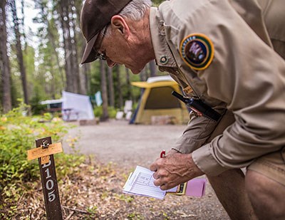 A man in a brown NPS volunteer uniform bends down to look at a numbered campsite post.