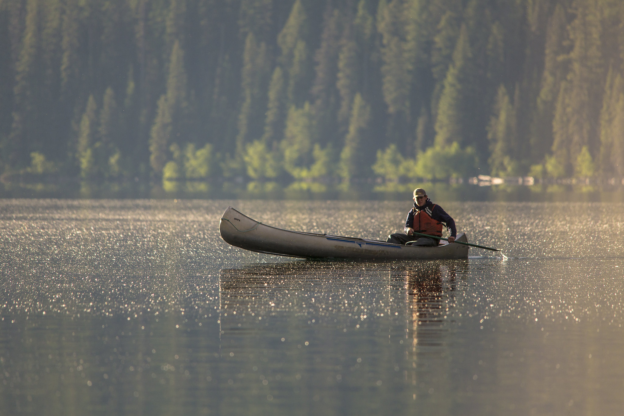 A man, in a canoe, paddles on a lake in the early morning sunlight.