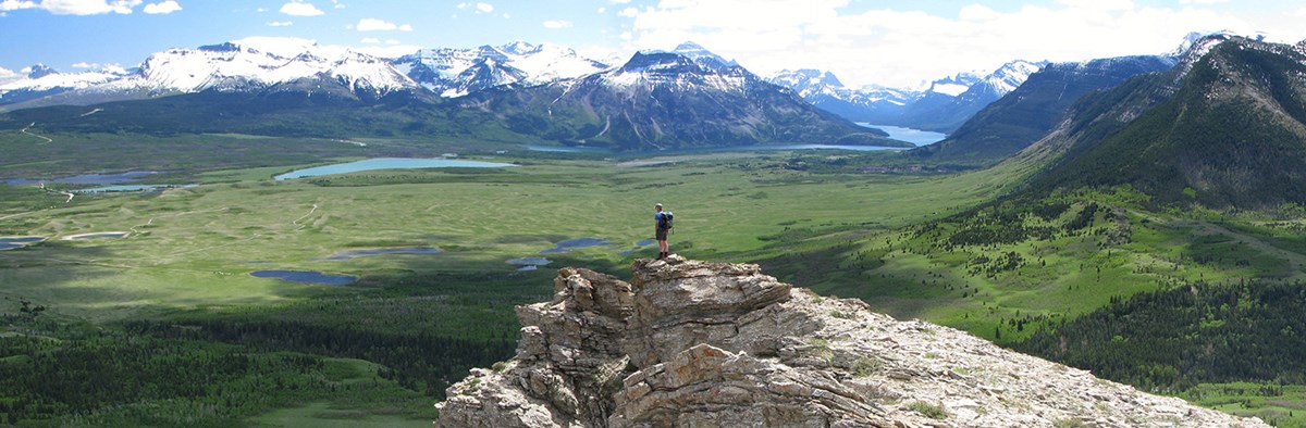 Hiker on craggy ridge looks out to where green expanse meets snowy mountain range