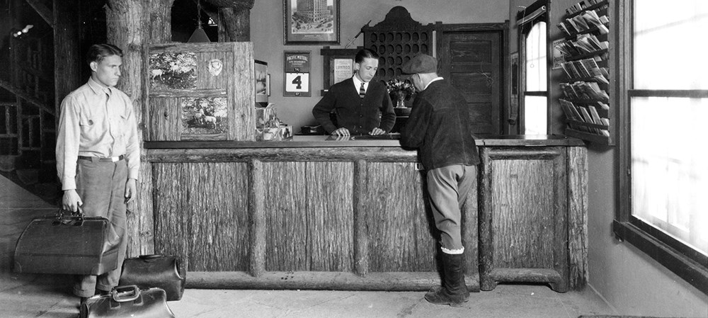 historic image of man at lodge reservation desk with employee holding luggage