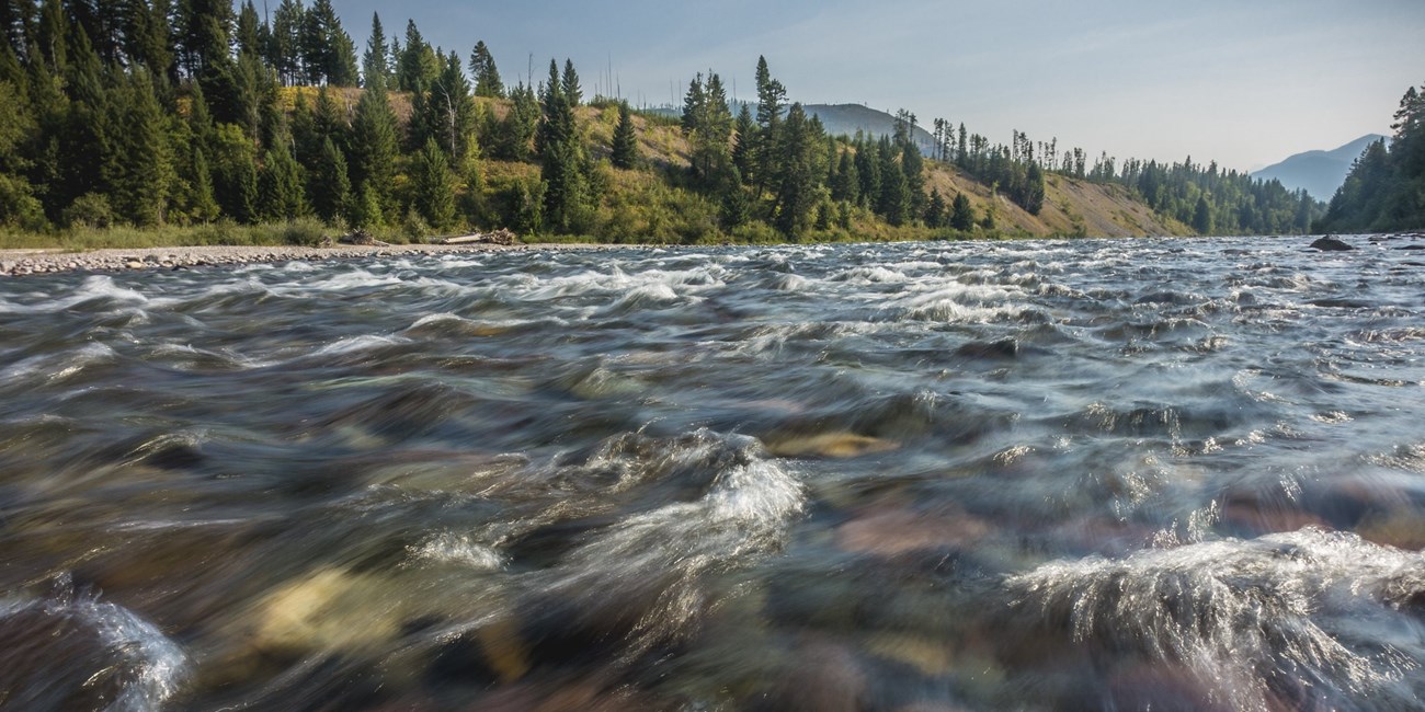 A river rushes in front of hills and trees.