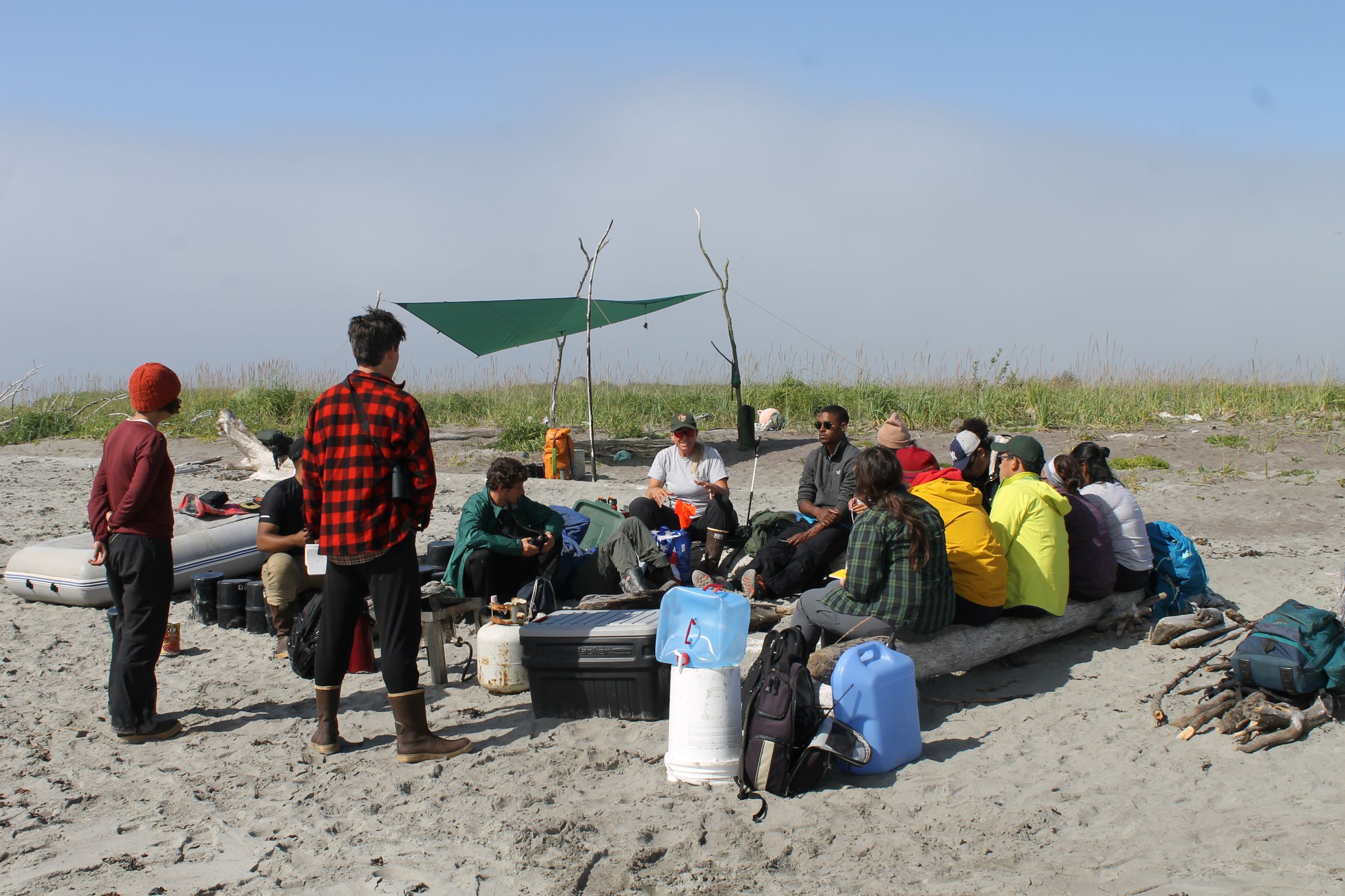 College students and instructors sitting on logs at a field camp with a tarp awning. 