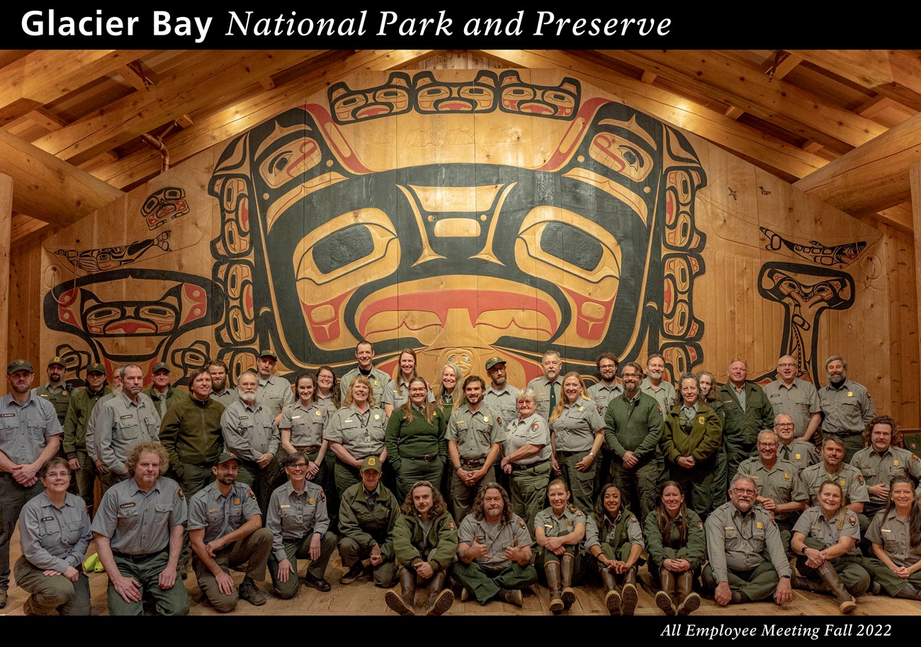 Glacier Bay staff Fall 2022. Several dozen staff members stand in front of the Huna Tribal House interior screen. Text reads Glacier Bay National Park and Preserve, all employee meeting fall 2022.