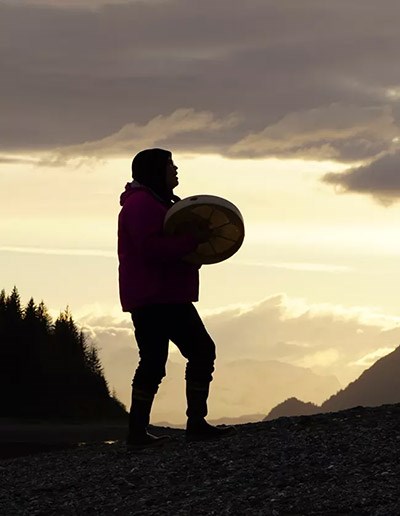 Silhouette of a Tlingit drummer against a bright sunset sky