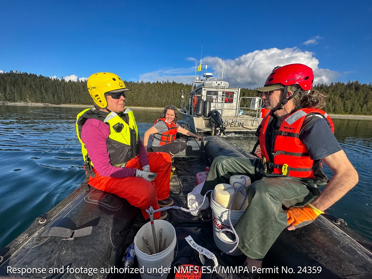 Three boaters wearing safety gear prepare to mobilize on their small vessel, a larger vessel, the Capelin, visible just behind them.