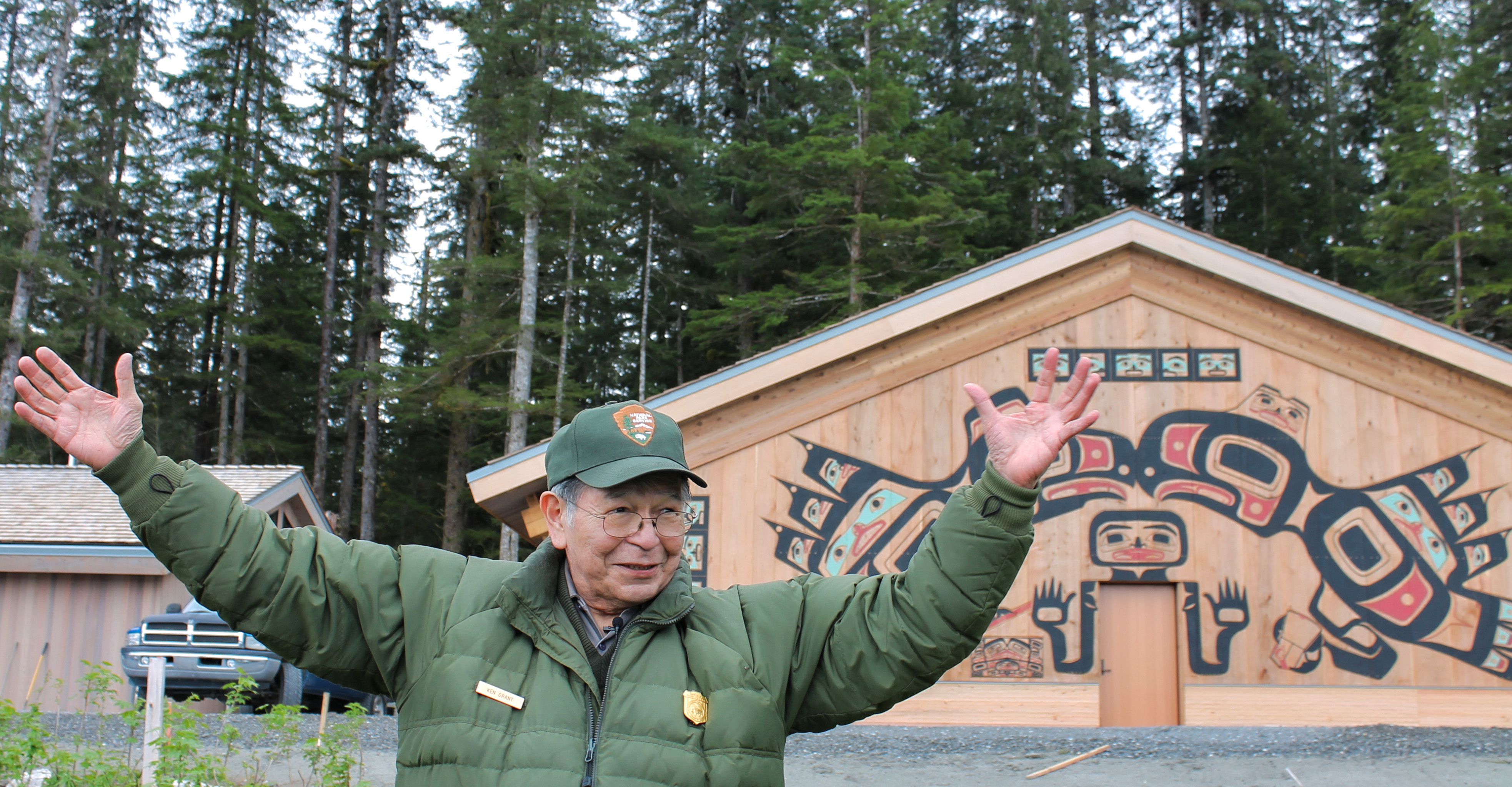 Elder Ken Grant smiles with arms outstretched standing in front of the Huna Tribal House in Glacier Bay.