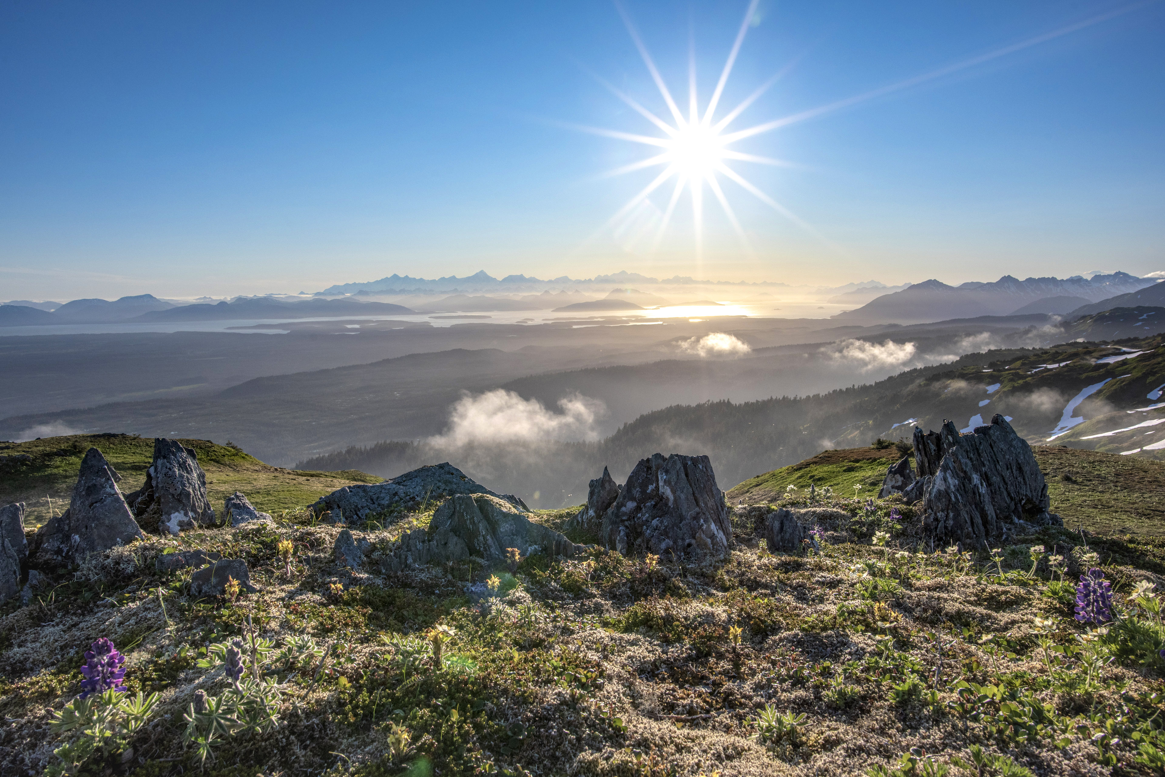 Vast view from atop a mountain ridge showing distant tall mountains, a bay of water and surrounding forested scenery. A bright sun shines in the blue sky.