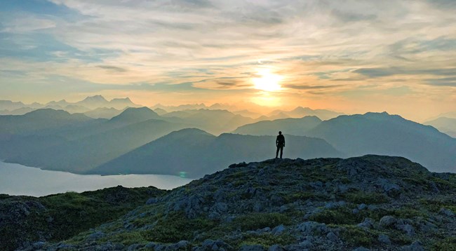 A lone silhouette of a person on a mountaintop overlooking a bay and rows of mountain ranges while a bright sun fills the sky.