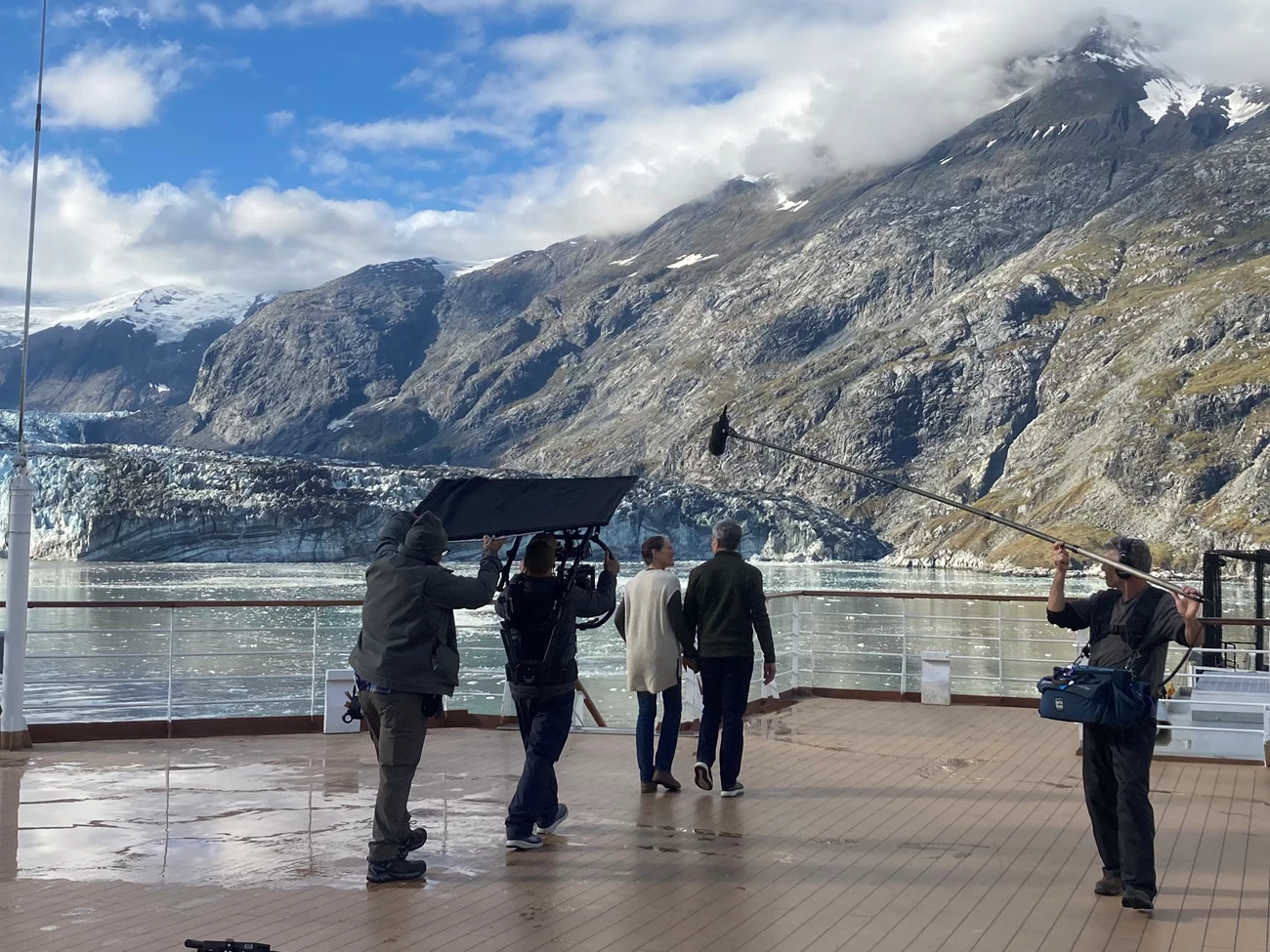 Commercial Filming on a cruise ship. A male actor and afemale actor walk hand in hand, while being filmed by a crew of three people. THey are on the deck of a cruise ship in front of a glacier with grey, snow peaked mountains in the background..