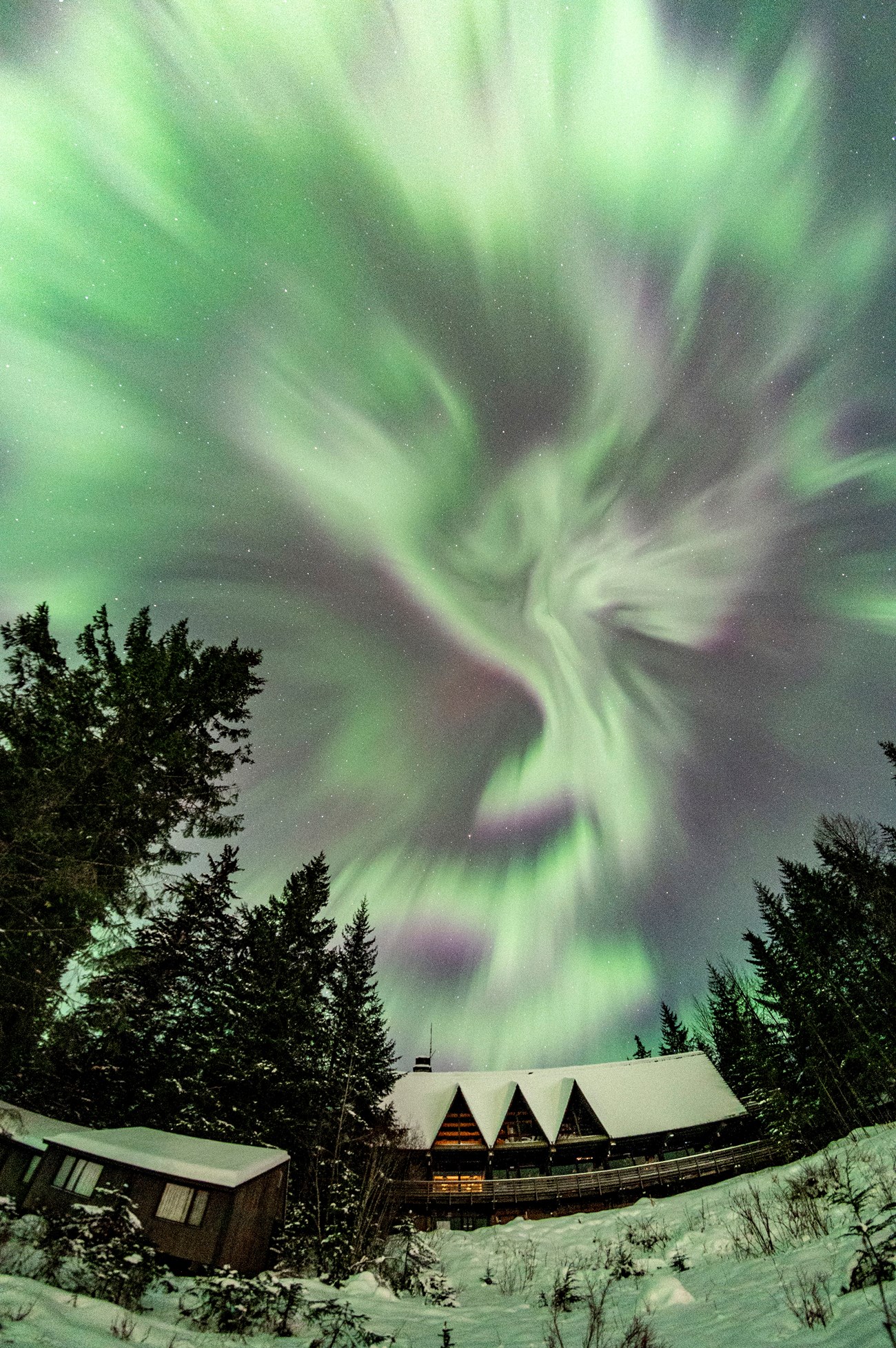 Green lights of the aurora borealis form spiraling abstract shapes in the sky above the snow-covered Glacier Bay Lodge.