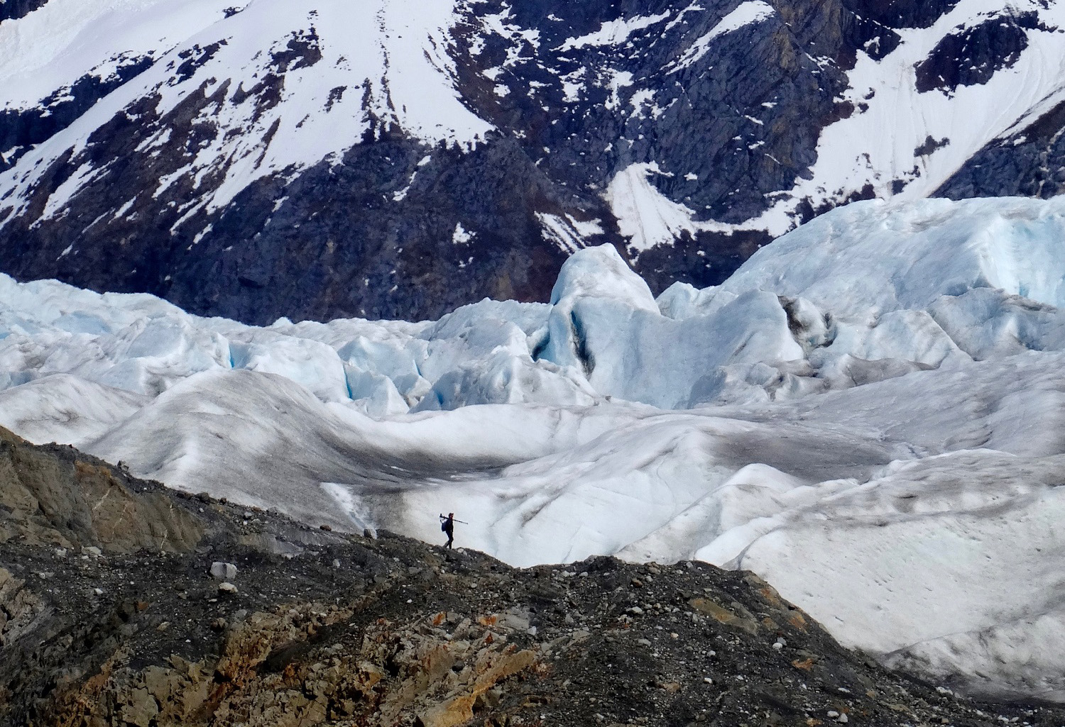 Silhouette of person walking with tripod dwarfed by glacier and mountain scenery behind.