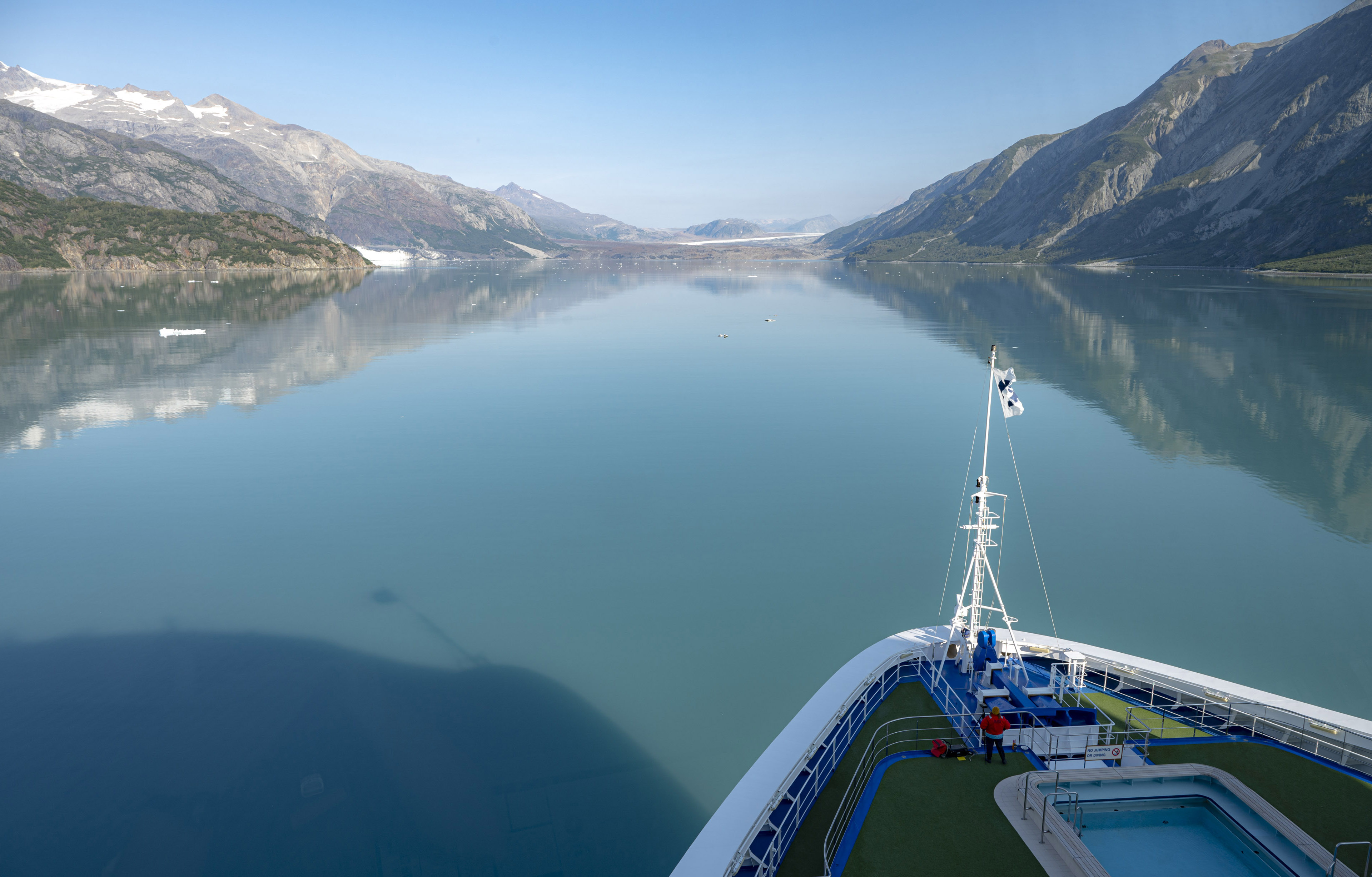 The bow of a cruise ship motoring through picturesque green water in a large valley. The shadow of the ship's bow is projected on the water on this blue sky day.