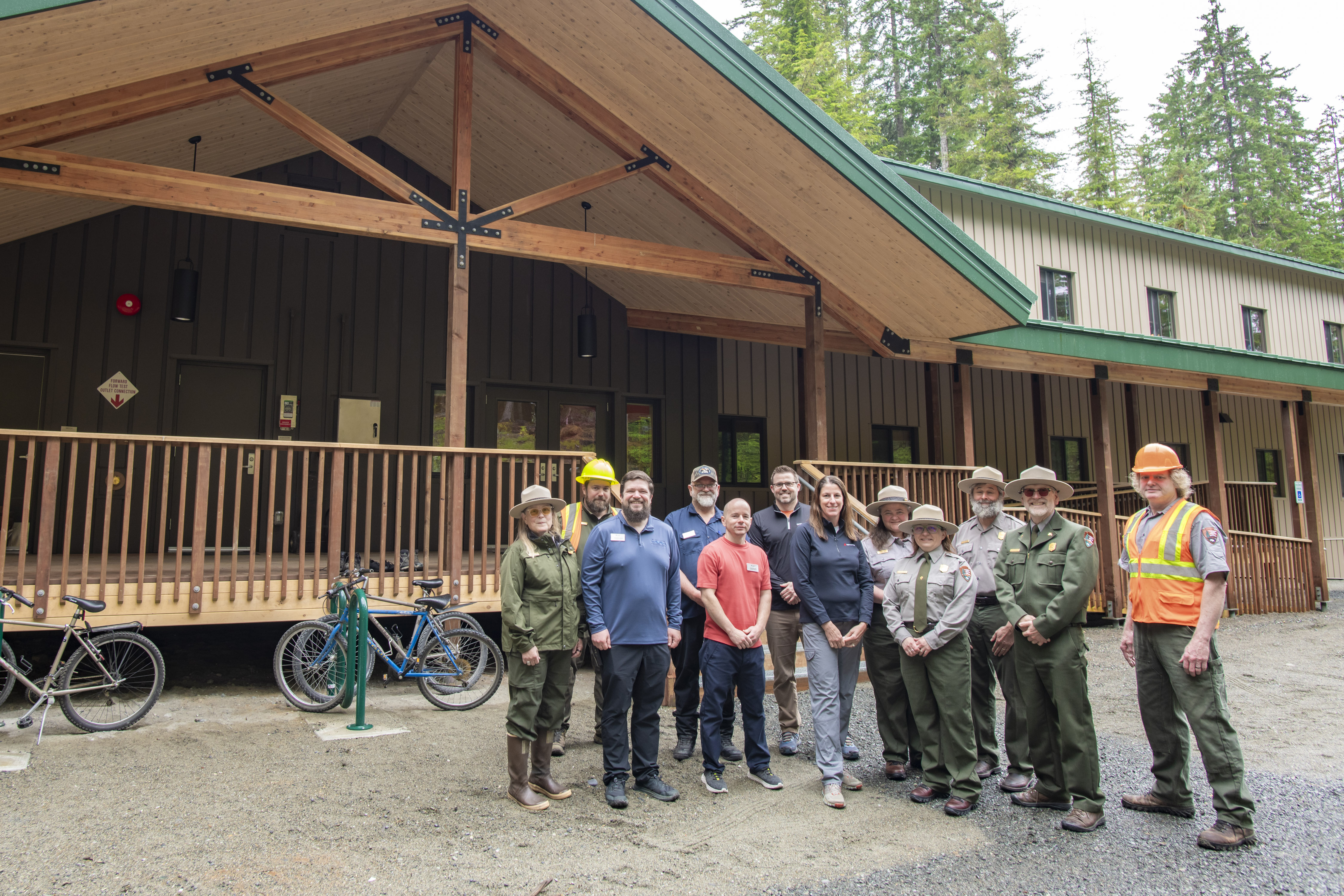 A group of twelve people stand in front of a newly constructed building, they stand in front of the staircase which leads to a covered entryway.