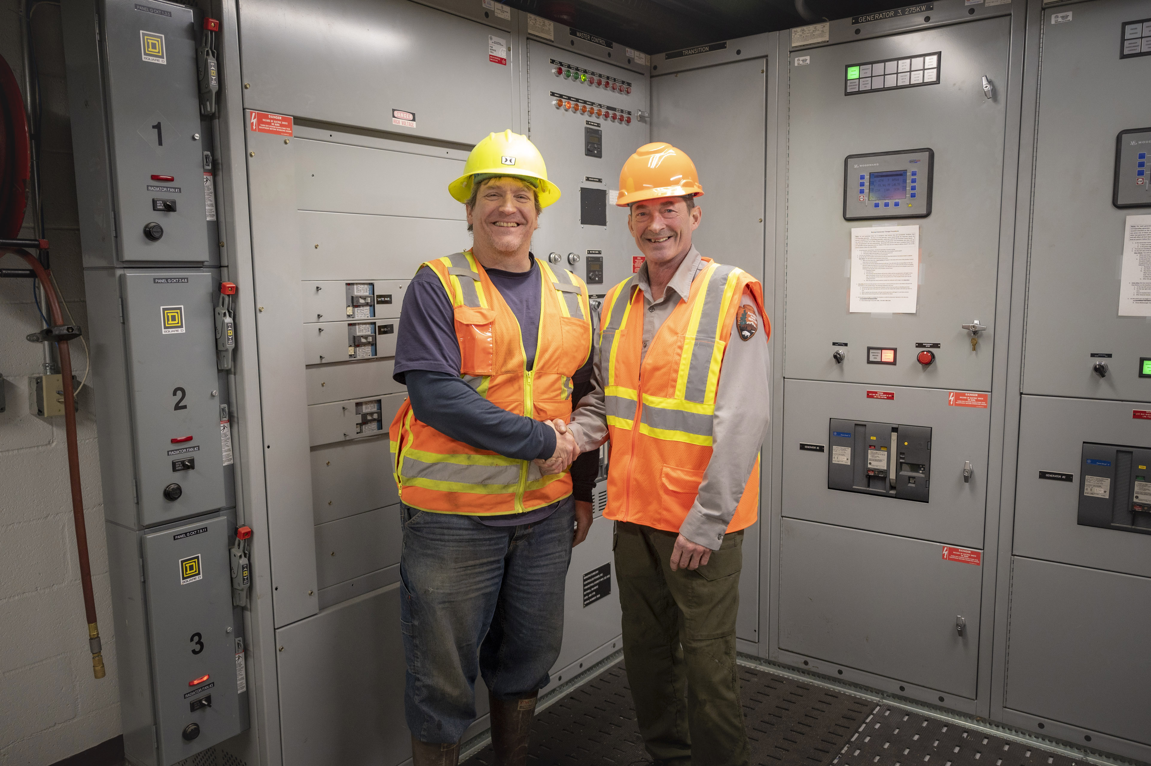 Glacier Bay Power Intertie Two people shake hands as they stand in safety vests and hard hats in front of electric panels and switches.