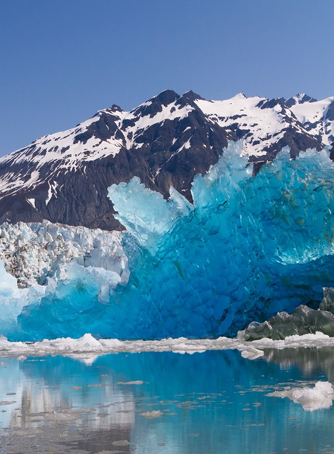 Rocky, snowy peaks frame the background of a water scene highlighted by a huge jagged chunk of electric blue ice, with a glacier visible behind it.