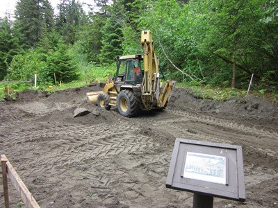 A backhoe works on a rectangular parcel of brown disturbed dirt. a sign in the foreground shows the future exhibit plan.