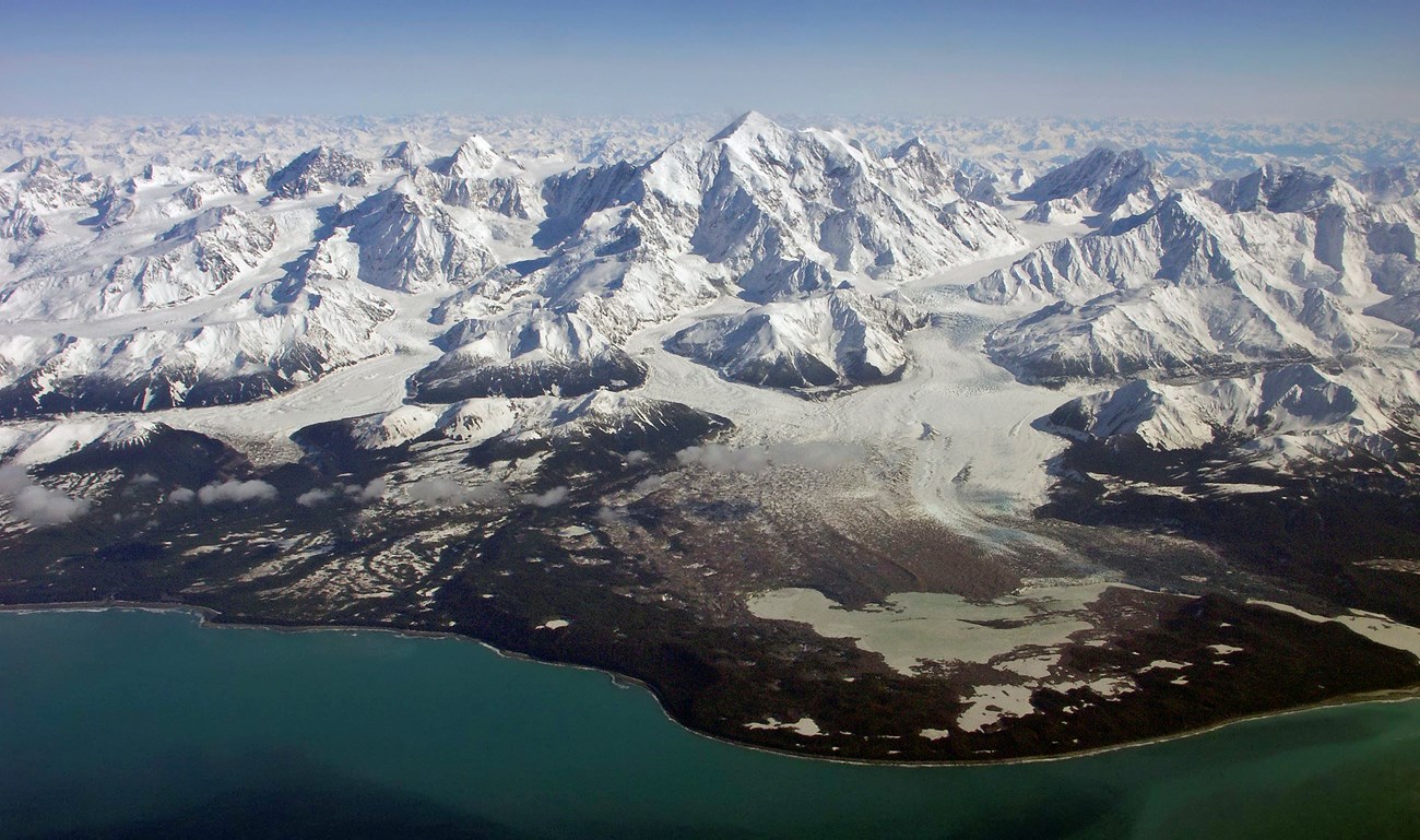Massive snowy mountains back a coastal aerial view. A glacier flows out of the central mountain, shaping the landscape.