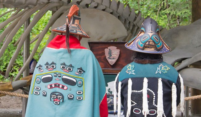 Two tribal members' backs face the camera, wearing decorative headwear, including formline art on a teal and red fabrics. The whale skeleton is out of focus in the background.