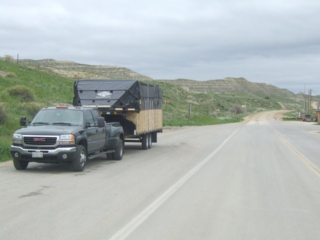 A heavy duty pickup truck is parked beside a 2-lane highway with small brown and grassy hills behind. The truck is pulling a large trailer.