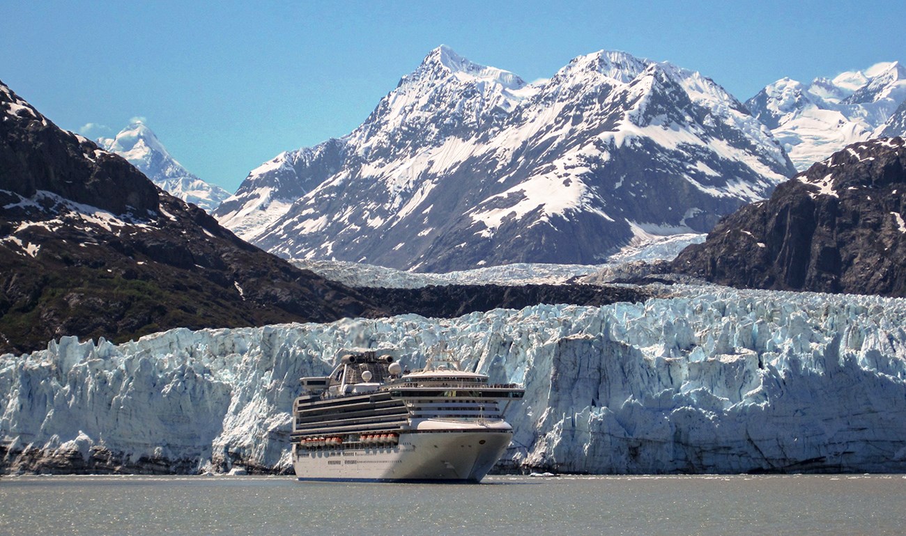 Huge cruise ship in brown-blue water with blue glacial ice and snowy mountains towering above