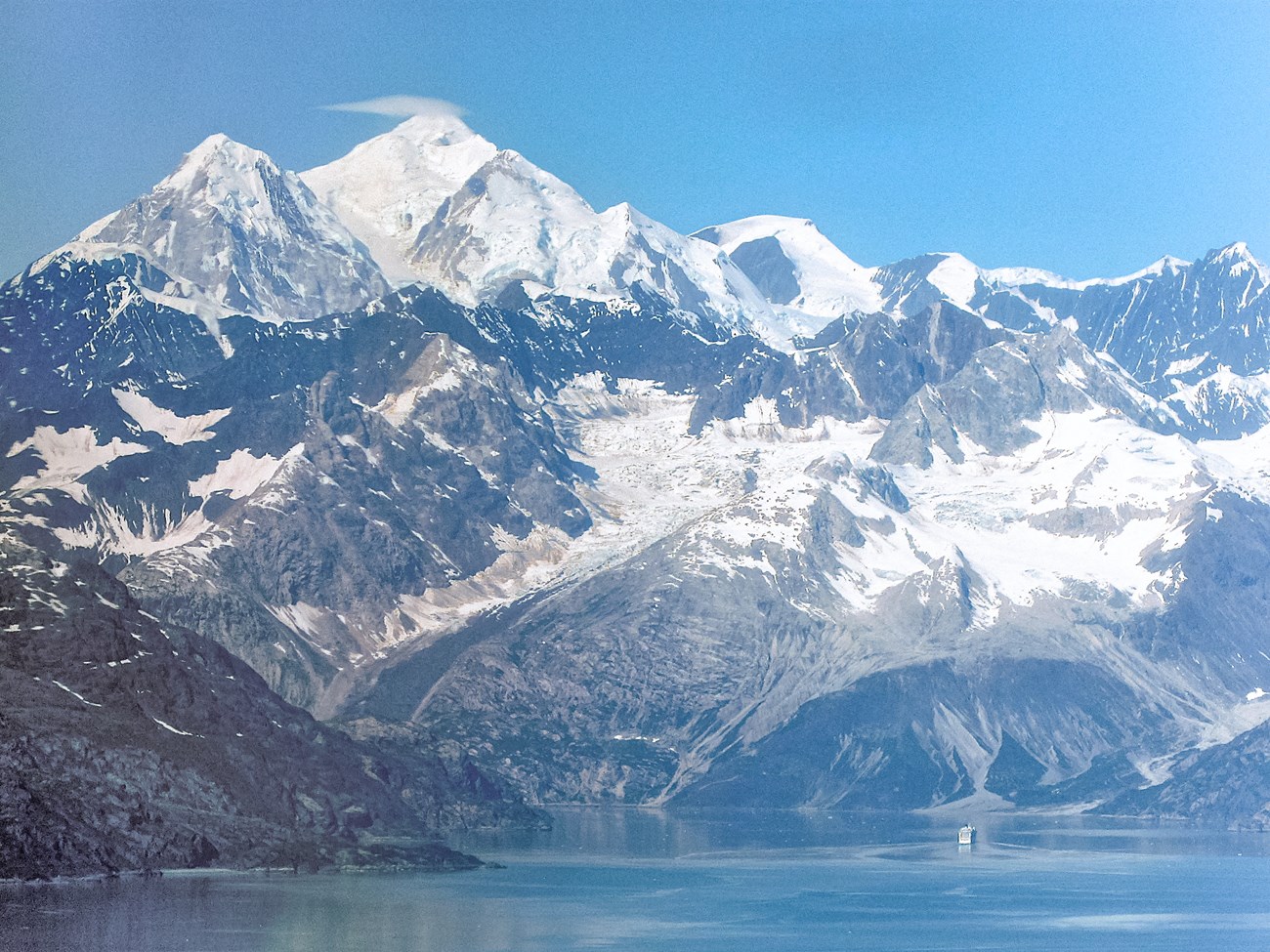 A cruise ship in glacier bay is dwarfed by mount fairweather rising thousands of feet above the ship.