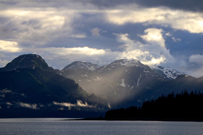 A wide mixture of clouds swirling high above mountains and hanging low over a waterway, the land covered in evergreen trees.